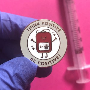 May include: A white enamel pin with a red cartoon blood bag with a smiling face and the text "Think Positive, Be Positive!" in gold lettering.