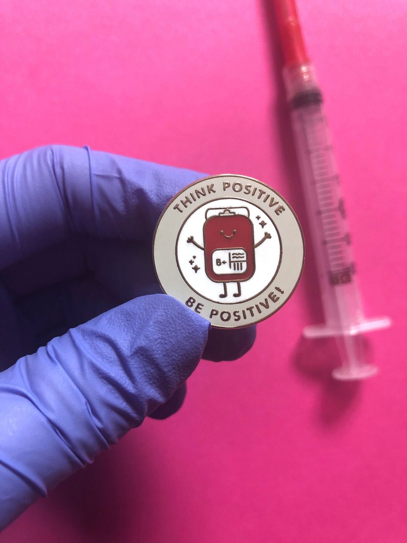 May include: A white enamel pin with a red cartoon blood bag with a smiling face and the text "Think Positive, Be Positive!" in gold lettering.