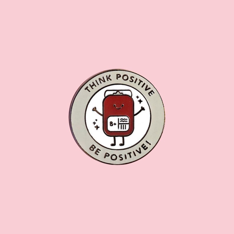 May include: A round pin with a white background and a red cartoon blood bag with a happy face. The blood bag has a label that says "B+". The pin has a gray border with the text "Think Positive" and "Be Positive!" around the edge.