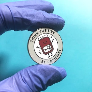 May include: A silver enamel pin with a white background and black outline. The pin features a cartoon blood bag with a smiling face and the text "Think Positive" and "Be Positive!" around the edge.