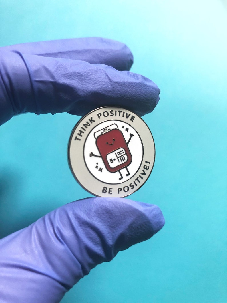 May include: A silver enamel pin with a white background and black outline. The pin features a cartoon blood bag with a smiling face and the text "Think Positive" and "Be Positive!" around the edge.