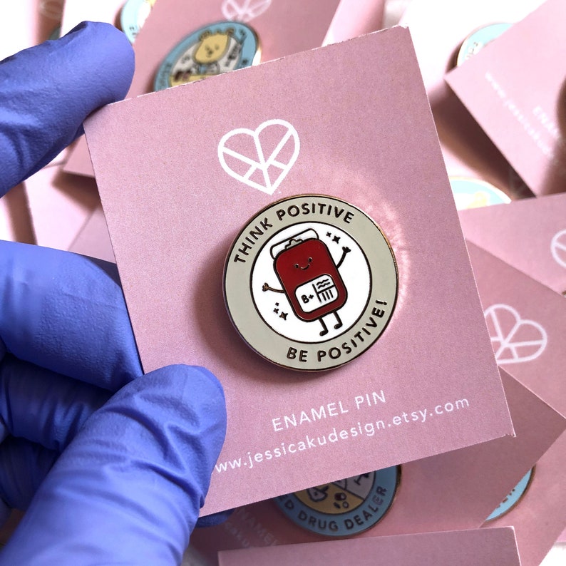 May include: A round enamel pin with a beige background and a red blood bag cartoon character with a smiling face and the text "Think Positive" and "Be Positive!" in white. The pin is on a pink card with the text "Enamel Pin" and a website address.
