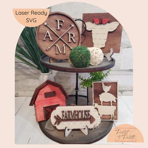 May include: A two-tiered wooden tray with farmhouse decor. The top tier features a round wooden sign with the word "FARM" and arrows, a wooden cow head sign, and a green moss ball. The bottom tier features a red wooden barn, a wooden sign with the words "FARMHOUSE" and arrows, and a wooden sign with a chicken, pig, and cow.