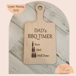 Op de afbeelding: Een houten snijplank met een handvat, gegraveerd met "Dad's BBQ Timer" en een grafische schaal van bierflessen om de gaarheid te meten: Rare, Medium, Well Done.
