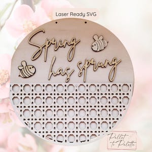 Pode incluir: Um sinal redondo de madeira com o texto "Spring has sprung" e duas ilustrações de abelhas. O sinal tem um padrão tecido na metade inferior.