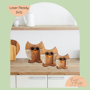 May include: Three wooden owl figurines of varying sizes, with brown bodies, black eyes, and orange beaks. The owls have decorative, light-colored wing patterns. The image includes the text "Laser Ready SVG" and "Pallet to Palette".