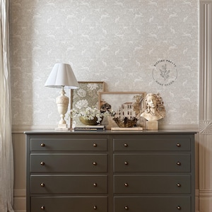 May include: A dark green dresser with six drawers and brass knobs. The dresser is against a wall with a white wallpaper featuring a repeating pattern of white rabbits. Above the dresser are three framed pictures and a white lamp with a white lampshade. The dresser is sitting on a wooden floor.