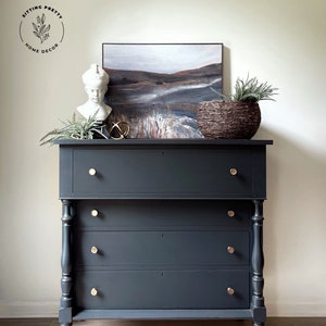 May include: A dark gray dresser with four drawers and gold knobs. The dresser has turned legs and is sitting on a light wood floor. There is a painting of a landscape above the dresser, a white bust, and a woven basket with a plant.