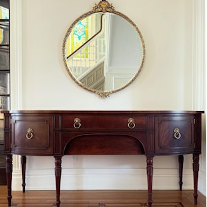 May include: A mahogany wood sideboard with a curved front and four legs. The sideboard has three drawers and two doors with brass lion head pulls. The sideboard is in front of a white wall with a round gold mirror above it.
