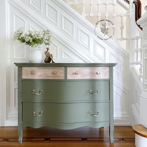 May include: A green painted dresser with three drawers and gold handles. The top two drawers have a wood veneer. The dresser is sitting on a wood floor with a white wainscoting wall behind it.