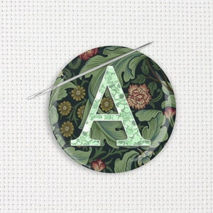 May include: A round, decorative pin with a green floral pattern and the letter "A" in a lighter green shade. A silver needle is placed across the pin. The pin features a dark background with flowers and leaves.