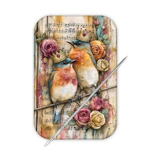 May include: A rectangular needle minder featuring two blue and orange birds wearing small gold crowns, perched on a branch against a background of vintage sheet music and colorful roses. A sharp metal sewing needle rests diagonally across the surface.