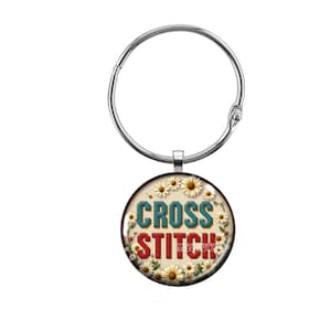 May include: A round, silver-colored charm with the words "CROSS STITCH" in teal and red, surrounded by daisies. The charm is attached to a silver-colored binder ring.