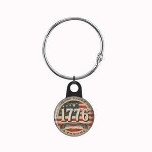May include: A circular metal keychain featuring a distressed American flag design with the number 1776 printed in large, bold, off-white font. The design is encased in a smooth, glossy finish and attached to a silver-toned metal ring.