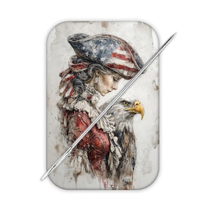 May include: A rectangular needle minder featuring a watercolor-style illustration of a person wearing a tricorn hat patterned with the American flag, standing beside a bald eagle. A long, thin metal sewing needle rests diagonally across the surface.