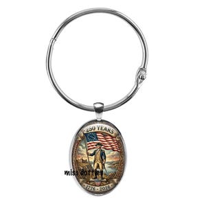 May include: A silver-colored metal ring with an oval pendant. The pendant features a historical illustration of a man holding a flag with the text "250 YEARS" and the dates "1776-2026". The flag has stars and stripes.