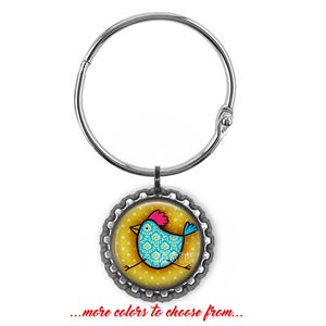 May include: A silver-toned metal ring attached to a circular bottle cap charm. The charm features a graphic of a blue chicken with a pink comb and tail on a yellow background with white polka dots. The text reads ...more colors to choose from...