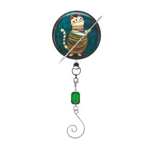 May include: A round, blue and green button with a white cat tangled in yarn. The button is attached to a silver chain with a green crystal bead and a silver hook.