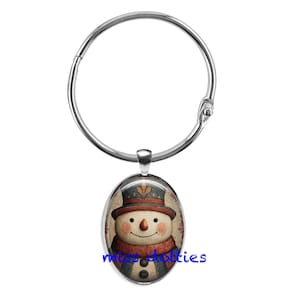 May include: A silver-colored metal ring with a decorative oval pendant. The pendant features a vintage-style snowman with a top hat, scarf, and buttons. The snowman has a carrot nose and a smiling face. The background is a light beige color.