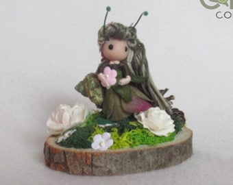 May include: A handmade fairy figurine with green and brown clothing, holding a pink flower. The fairy is standing on a wooden base with moss and white flowers. The base is not included.