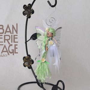 May include: A green and white fairy doll with a butterfly wing design, long green yarn hair, and a white feather skirt. The doll is hanging from a black metal stand with a butterfly design.