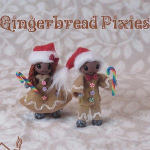 May include: Two handcrafted gingerbread pixie dolls. Each doll wears a red and white Santa hat and holds a rainbow candy cane. The dolls are dressed in gingerbread-themed outfits with colourful accents. The text "Gingerbread Pixies" is at the top.