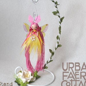 May include: A handmade fairy doll with pink and yellow yarn hair, a yellow and white dress, and white wings. The fairy is hanging from a white metal stand with green leaves.