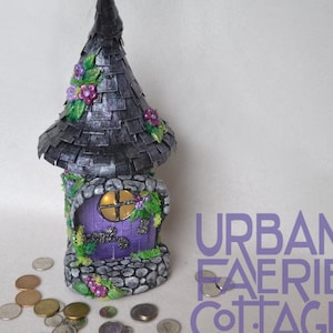 May include: A purple and gray fairy house with a pointed roof and a small door. The house is decorated with flowers and vines. Coins are scattered around the house. The text "URBAN FAERIE COTTAGE COMPANY" is visible in the lower right corner of the image.