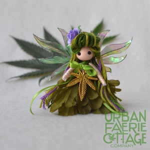 May include: A green fairy doll with iridescent wings, green yarn hair, a purple flower, and a gold marijuana leaf pendant. The doll is wearing a green dress with purple trim.