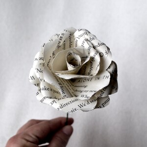 Puede incluir: Una rosa blanca de papel hecha con las p&aacute;ginas de un libro. La rosa est&aacute; enroscada y tiene un tallo verde. El texto en las p&aacute;ginas es visible, incluyendo las palabras "Alone in the" y "bring my new".