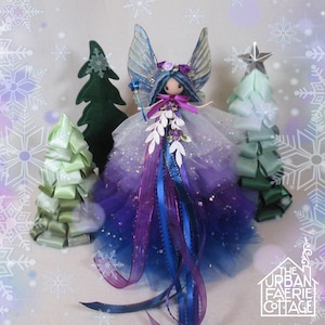 May include: A handmade fairy doll with a purple and blue ombre tulle dress, blue wings, and a wand. The fairy is surrounded by decorative green and silver Christmas trees. The Urban Faerie Cottage logo is in the bottom right corner.