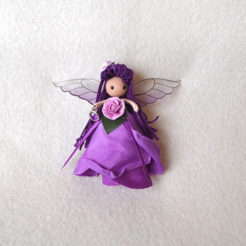 May include: Purple fairy doll with wings, a purple flower, and a purple gem. The doll is made of fabric and yarn and has a wooden head.
