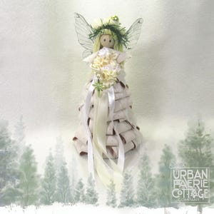 May include: A white fairy doll with green wings and a white flower crown. The doll is wearing a white dress made of layered fabric and ribbon. The doll is standing on a white background with a green watercolor background.