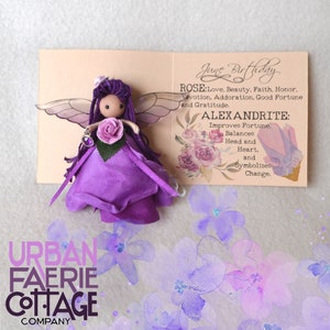 May include: A purple fairy doll with wings, a purple dress, and a pink rose in her hair. The doll is sitting on a card with the text "June Birthday" and information about the rose and alexandrite birthstones.