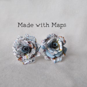Puede incluir: Dos rosas de papel hechas con mapas antiguos. Las rosas son de color azul claro y blanco con un centro gris claro. El texto "Made with Maps" est&aacute; por encima de las rosas.