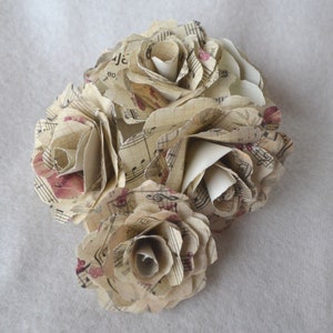 Puede incluir: Un ramo de cuatro rosas de papel hechas con partituras musicales antiguas. Las rosas son de color crema con acentos rojos y est&aacute;n dispuestas en un racimo.