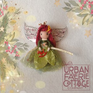 May include: A handcrafted Christmas angel doll with red yarn hair, a gold crown, and glittery wings. The doll is dressed in a green gown and holds a red rocking horse. The background features a watercolour design with stars and foliage.