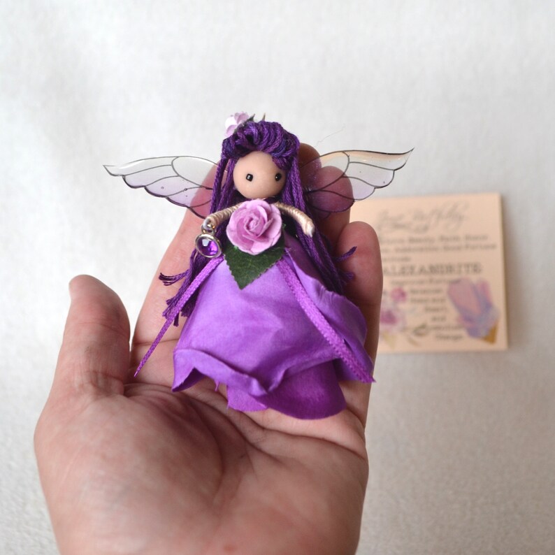 May include: Purple fairy doll with wings, a purple dress, and a flower crown. The doll is holding a small purple gem.