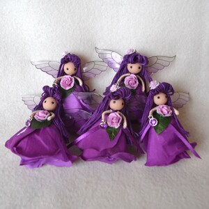 May include: Five purple fairy dolls with wings, each holding a purple rose and wearing a purple dress. The dolls are made of fabric and have yarn hair.