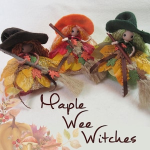 Puede incluir: Tres muñecas de brujas hechas a mano con faldas de hojas, sombreros y escobas. Las muñecas tienen sombreros y pelo naranja, marrón y verde. El texto "Maple Wee Witches" está escrito en marrón. Tema otoñal.