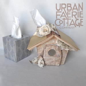 May include: Two tissue boxes, one is a plain white box with a tissue pulled out, the other is a book-shaped tissue box with a brown cover and a white tissue pulled out. The book-shaped tissue box is decorated with paper flowers and the text "URBAN FAERIE COTTAGE COMPANY".