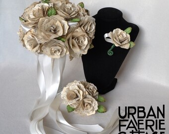May include: A bouquet of cream-colored paper roses made from book pages, with white satin ribbon streamers. A matching boutonniere is displayed on a black stand. The text "URBAN FAERIE COTTAGE COMPANY" is visible below the bouquet.