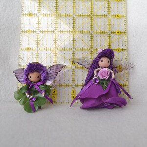 May include: Two purple fairy dolls with wings and flower accents. One fairy is holding a purple flower and the other is holding a shamrock.