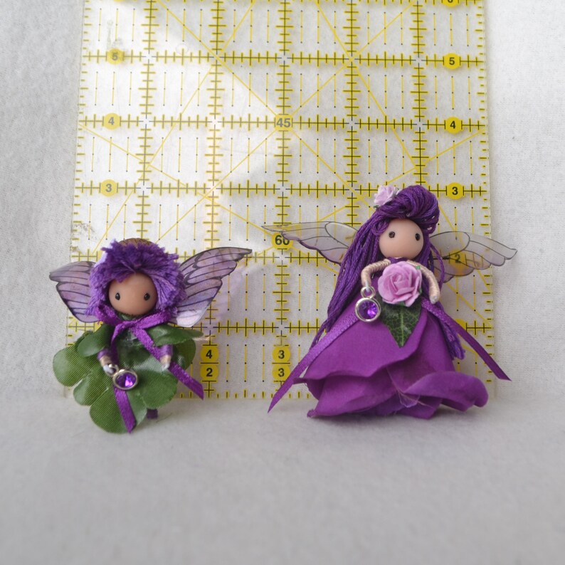 May include: Two purple fairy dolls with wings and flower accents. One fairy is holding a purple flower and the other is holding a shamrock.
