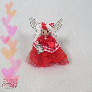 May include: A handcrafted fairy doll with white hair, a red and white checkered dress, and delicate wings. The doll holds a small book-shaped charm that reads "Once Upon a Time". The fairy has a red bow and a pearl necklace.