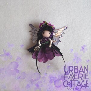 May include: Purple fairy doll with black hair, a purple flower crown, and wings. She is wearing a purple dress and holding a purple flower. The doll is on a white background with purple flowers.