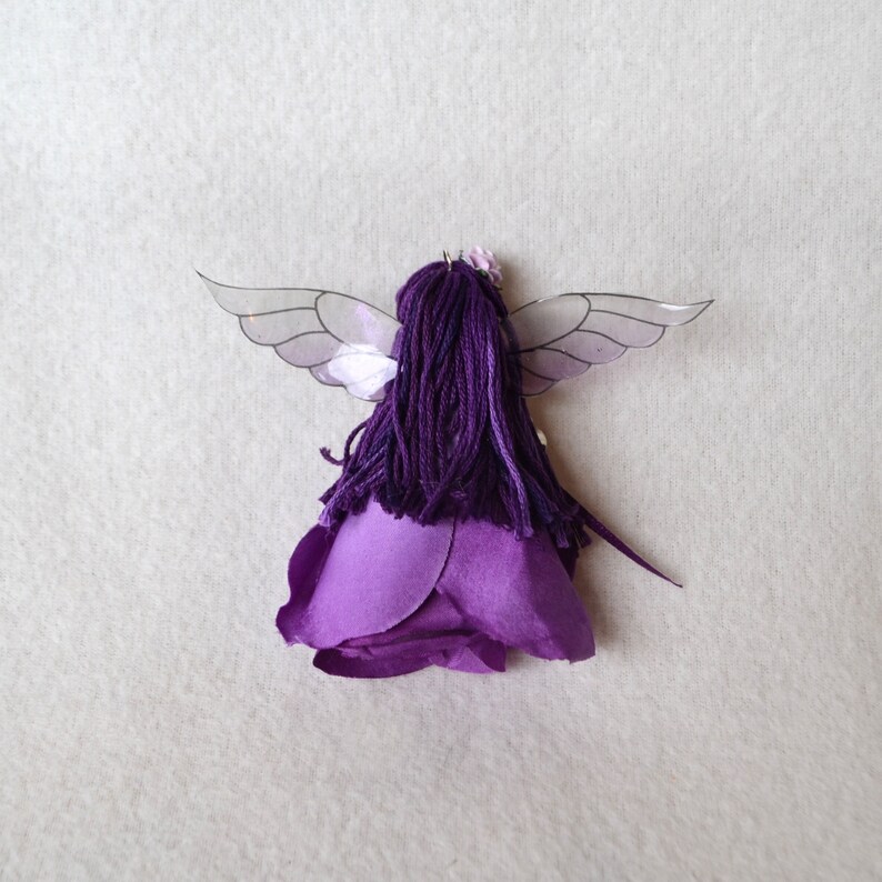 May include: Purple fairy doll with wings made of clear plastic and a purple flower crown. The doll is wearing a purple dress made of fabric that resembles a rose.