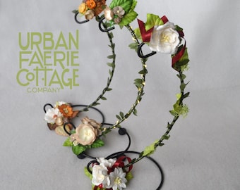 Puede incluir: Tres diademas florales con hojas verdes y flores blancas, rojas y naranjas sobre un soporte de metal negro. Las diademas están decoradas con cinta roja y hojas verdes. El soporte tiene una base circular y dos brazos curvos.