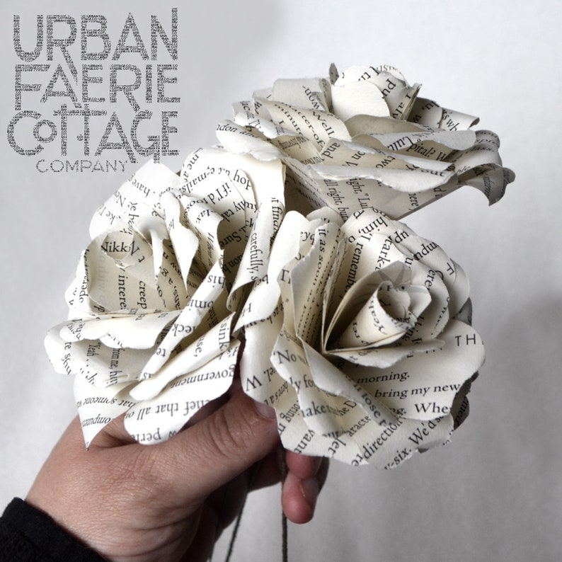 Puede incluir: Tres rosas blancas de papel hechas con p&aacute;ginas de libros. Las rosas se sostienen en una mano. El texto "URBAN FAERIE COTTAGE COMPANY" es visible en la parte superior de la imagen.