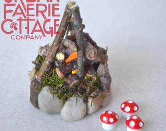 Può includere: Un falò in miniatura fatto di ramoscelli e pietre con una luce arancione incandescente all'interno. Tre figurine di funghi rossi e bianchi sono in primo piano.